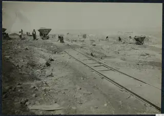 Excavation at Vouni with narrow rails, two tipping wagons and workers among low stone walls and rubble.