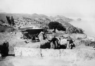 Four people digging with spades at the Vouni excavation, with a rail tipping wagon at left and the sea beyond.