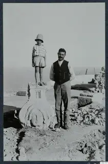 A boy stands on a carved limestone sculpture with a face, and a man stands beside it among excavated ruins at Vouni.