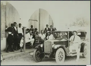 Group of men and a small child posing outside an arched doorway beside an open-top car with a tall radiator, in Pyrgos