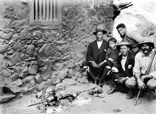 Five people sit and stand by a stone wall, roasting a whole goat on a spit over a fire at Galini.