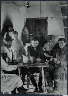 Nine men gather around a small wooden table playing cards in a whitewashed room with a wooden door.