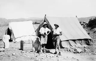Two men in pith helmets stand outside canvas tents at Vouni, with a stone sculpture fragment at the left edge.