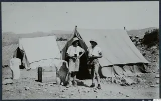 Two men in pith helmets stand outside large canvas tents at Vouni, with a stone sculpture fragment at left.