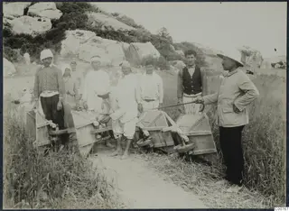Workers with two wooden wheelbarrows on a grassy path at Oura, with a man in a hat standing to the right.