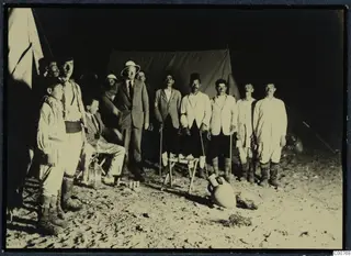 Twelve men posed outside tents at night, with a large amphora on the sand in front.
