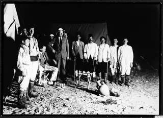 Men standing and seated outside dark tents on sand in low light, with a large two-handled amphora on the ground in front.