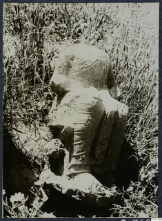 Weathered limestone statue of a figure, partly buried in tall grass at Oura.