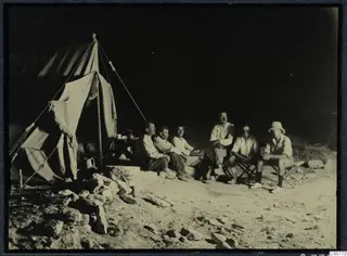 Six men sitting outside a canvas tent on sand at night, one at the right wearing a pith helmet.