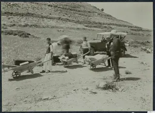 Men with wheelbarrows loaded with boxes and bundles on a dry hillside, with an open car behind them.