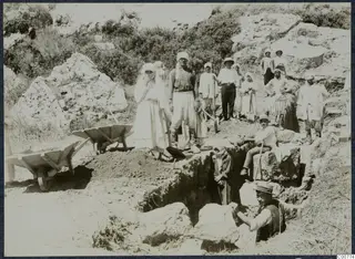 Workers with shovels beside a trench and two wheelbarrows during excavation at Oura