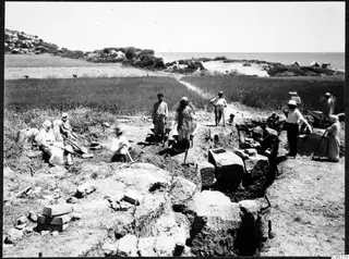 Workers with spades and hoes at an excavation of large stone blocks in a field, with the sea in the background.
