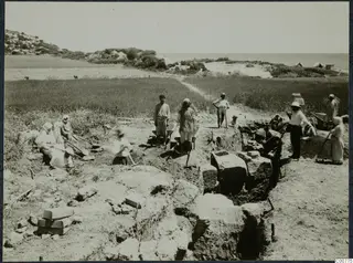 Workers with spades and hoes at an excavated trench among stone blocks, with a green field and the sea behind.