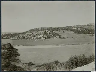 Rocky hills with scrub and open grassy fields near Oura, with the sea visible on the horizon at left.