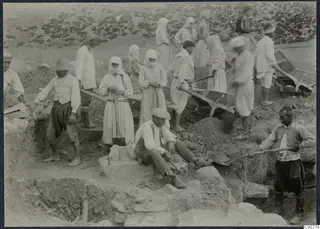 Workers digging at Oura, with wheelbarrows and shovels among stone walls in an excavation trench.