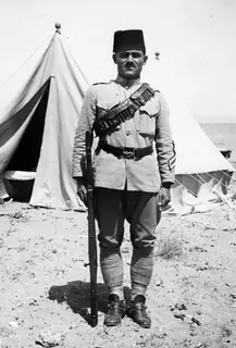 Standing policeman in uniform and fez with rifle and ammunition bandolier, in front of tents on sand, at Oura.