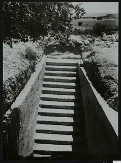 Stepped dromos cut down between walls toward a tomb entrance at Trachonas, with the sea in the distance.