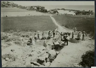 Excavation at Oura with about eighteen workers, wheelbarrows and a spoil heap, with the sea behind