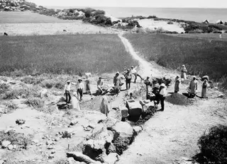 Workers at the Oura excavation with wheelbarrows, standing and sitting along a trench edge, with the sea in the distance