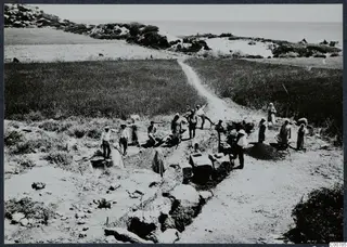 Workers at the Oura excavation, with a trench, exposed stone blocks and wall foundations, beside cultivated fields and the sea.