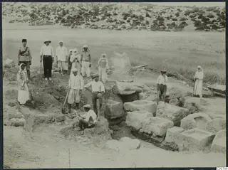 Workers and a uniformed man standing and kneeling among cut stone blocks at an excavation.