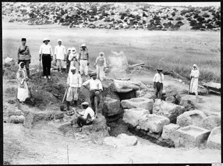 Workers and local people gathered at an excavation among cut stone blocks, with a uniformed man standing at left.