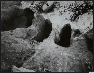 Excavation at Lapithos Kastros showing two dark arched openings cut into the soil, with a stone rubble wall above.