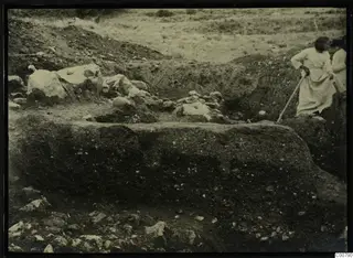 Excavation trench at Plakes, Lapithos, with two people working beside stones and a long-handled tool.