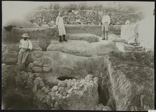 Excavation at Plakes, Lapithos, with three people standing and sitting among cut earth terraces and stone walls.