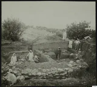 Excavation at Plakes, Lapithos, with workers in a trench beside stone walls, and a wheelbarrow on the bank at right.