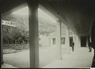 Two men stand in a colonnade with a tiled walkway, with a rocky mountain and a white building behind the courtyard