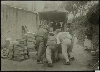 Men loading stacked boxes onto a covered cart in a dirt lane at Lapithos, with onlookers behind