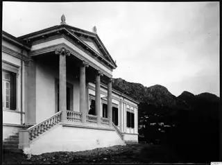 Columned building with a pediment and akroterion, a man on its upper terrace, mountains rising behind
