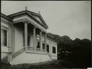 Building with a columned porch and pediment, a man standing on the terrace, mountains rising behind