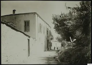 Whitewashed two-storey building beside a dirt lane at Lapithos, with trees on the right and two people on the track