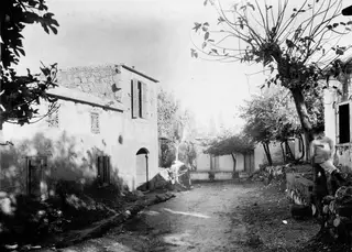 Dirt street in the village of Lapithos lined with whitewashed houses, trees and low stone walls, with one man leaning against a tree.