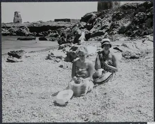 Two women sitting wrapped in towels on a pebble beach, with rocks and the sea behind them at Lambousa
