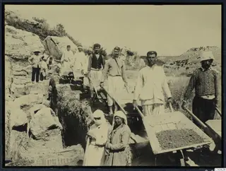Workers with wheelbarrows and shovels in an excavation trench cut between rock outcrops, with a row of men on the left.