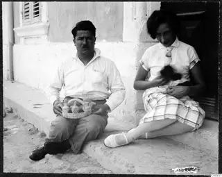 Two people seated on a doorstep outside a whitewashed house; the man holds a small handled tray of little vessels.