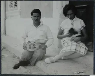 A man and a woman sitting on a whitewashed doorstep; he holds a tray of small vessels and she holds a cat.