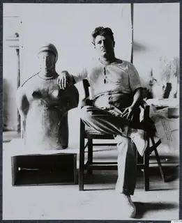 Toulis Souidos seated beside a large sculpture of a figure with a beaded necklace and cap
