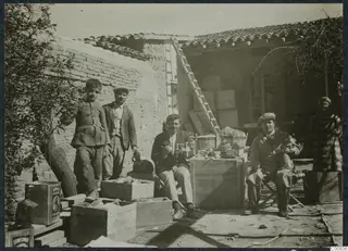 Courtyard with five people among wooden crates, jugs and vessels, a ladder leaning on a wall and a tiled-roof building behind