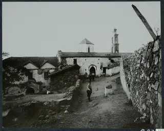 Monastery at Machairas seen from a dirt track, with whitewashed walls, tiled roofs, a bell tower and three people