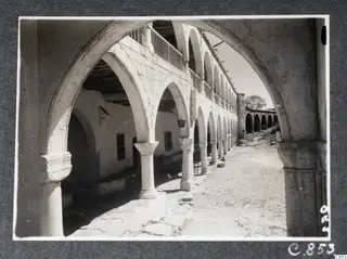 Arcaded courtyard at Lambousa with stone arches, columns with carved capitals, and an upper balcony railing.