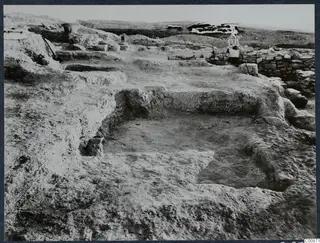 Excavated square pits at Idalion with a low stone wall and a seated person beyond, seen across the dig