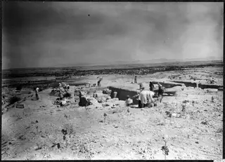 Workers excavating walls and trenches on the top of the acropolis at Idalion, with a wheelbarrow and the plain below.