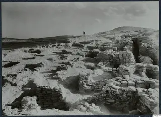 Excavated stone walls and foundations at Idalion Acropolis, with two people on the ridge and bare hills behind.