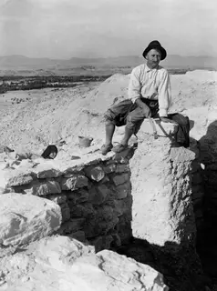 Panajiotis sitting on a stone block at the excavation at Idalion, holding a tool, with a head in a dark headscarf behind the wall.
