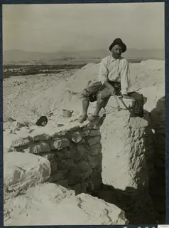Panajiotis sitting on a stone wall at the excavation at Idalion, with hills and a valley behind.