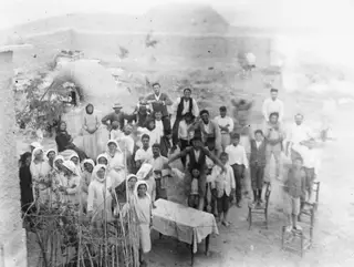 Farewell party at Panajiotis, Idalion: a crowd posed in a yard by a mud-brick wall, with a table and chairs.